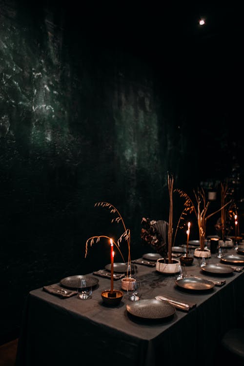 Gloomy banquet table with dark tableware and tablecloth decorated with compositions of dry branches and stems and burning candles in dark room