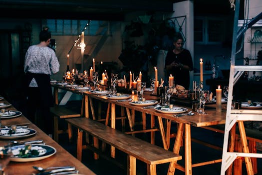 Anonymous women serving wooden tables with plates and glassware among burning candles while preparing for banquet in modern place