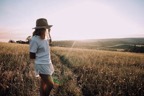 Side view of anonymous woman in white summer outfit and hat standing in field on sunny day