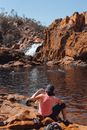 Side view of full body anonymous female traveler in sportswear and hat sitting on brown rocky coast and admiring picturesque landscape with small waterfall and swimming people on summer day