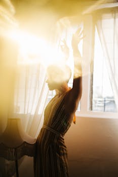 A woman in a striped dress gracefully raises her arms in a sunlit room, exuding peace and inspiration.