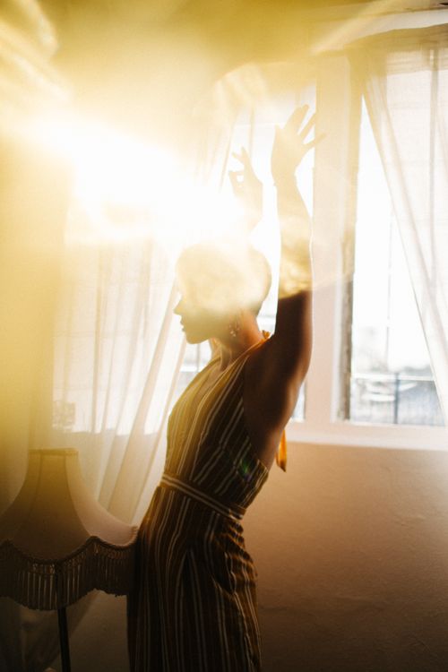 Free Young anonymous female with arms raised in striped dress near window in bright light of sun Stock Photo