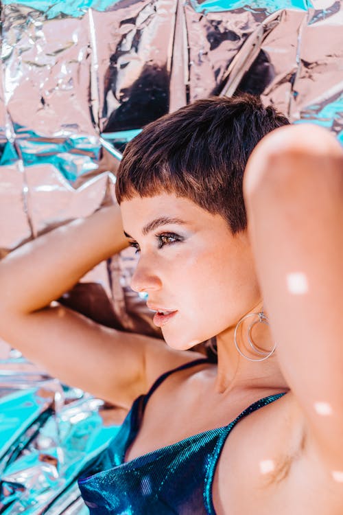 Side view of self assured young female in trendy outfit touching short dark hair and looking away against reflecting foil background