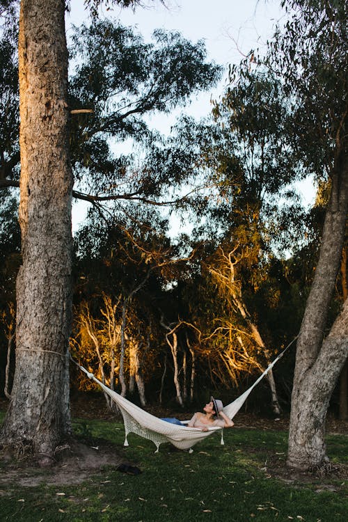 Unrecognizable female traveler in casual clothe and hat lying in hammock in green forest in evening
