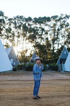 Side view of young trendy female traveler in stylish denim outfit and hat talking on mobile phone while standing near small similar houses in nature