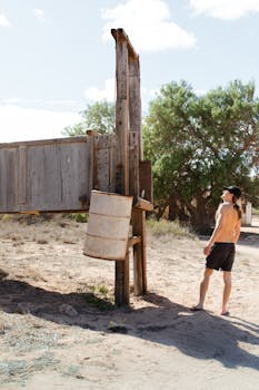 Back view of unrecognizable shirtless male traveler with long hair in shorts and cap standing on sandy terrain near old wooden fence and looking up