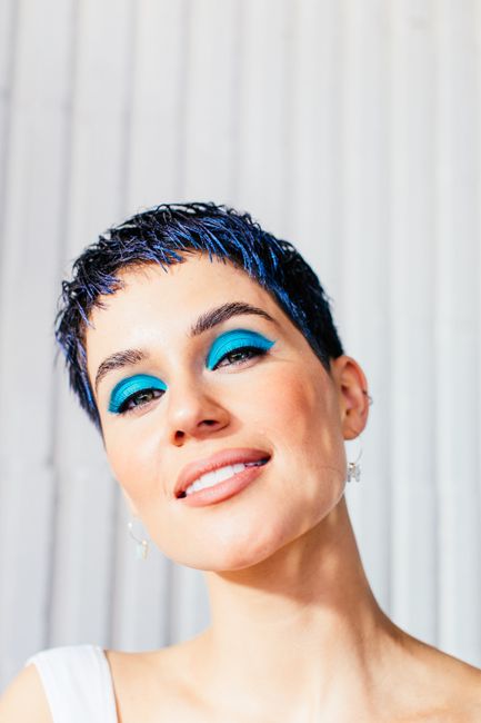 Delighted young female model with short dark hair and blue eyeshadows smiling and looking at camera in light studio