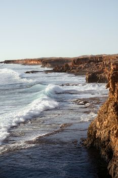 汹涌的海浪拍打着海岸，岛上巨石嶙峋的海岸线在万里无云的天空下延伸