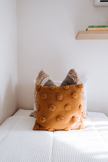 Interior of bedroom with soft cushions placed on bed with white blanket and stack of books on shelf hanging on wall