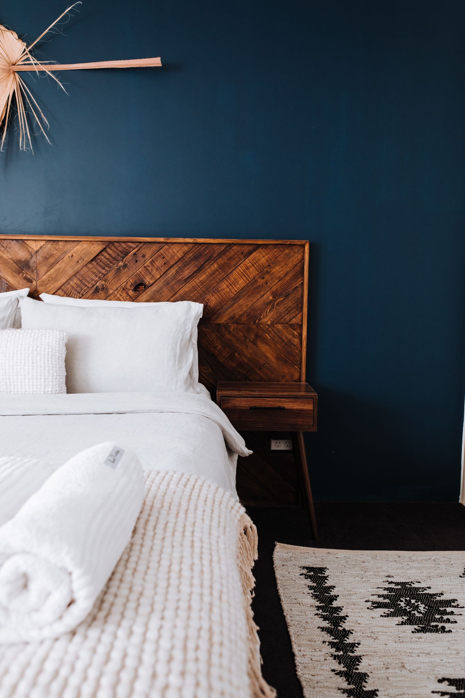 Deep blue bedroom wall behind a warm wooden headboard and white bedding.