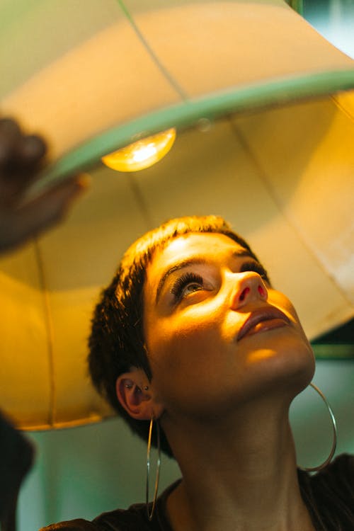 Stylish female with makeup wearing earrings under lamp with shining light bulb looking up