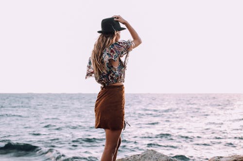 Free Back view of anonymous female with hand on head in stylish outfit standing on coast and admiring rippling sea Stock Photo
