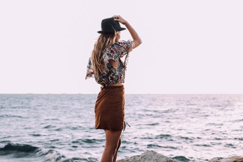 Back view of anonymous female with hand on head in stylish outfit standing on coast and admiring rippling sea