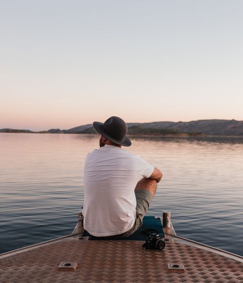 Free Back view of unrecognizable male tourist with photo camera sitting on quay near calm rippling river at sundown Stock Photo