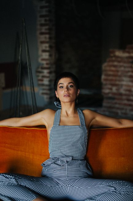 Trendy serious female with short dark hair wearing checkered overall looking at camera while sitting on couch with bent legs in room with brick interior