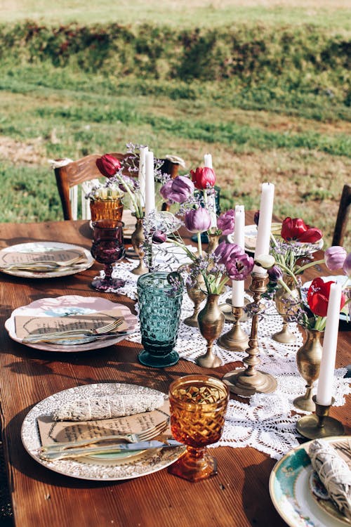 Table setting with plates and glassware placed near candles and flowers placed on grassy field in countryside during holiday celebration