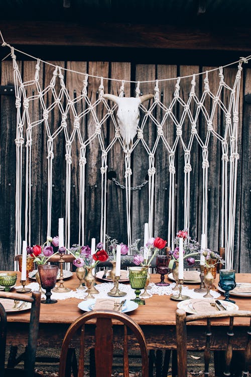 Festive table setting with glassware and plates placed near candles and colorful flowers on terrace with skull on wooden wall