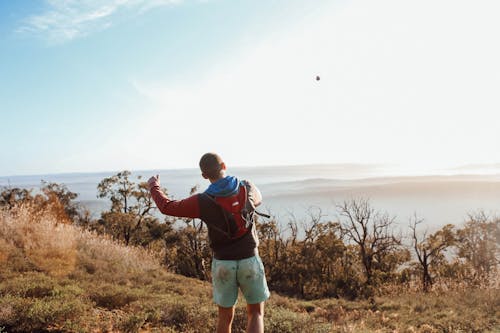 Back view of anonymous male explorer throwing stone while standing on grassy hilltop above foggy mountain peaks in sunny nature