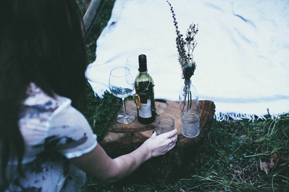 Free From above of unrecognizable female wineglasses and bottle of alcohol drink on grassy ground with blanket during picnic in nature Stock Photo