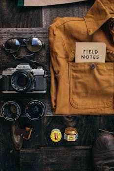Flat lay of vintage camera gear, field notes, sunglasses, and accessories on a rustic table.