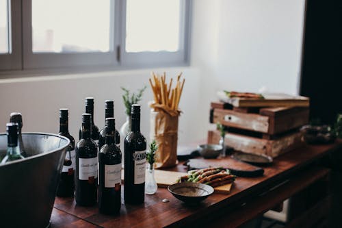 Various bottles of alcoholic drinks placed on table with grissini sticks and wooden boxes near green branches in vase in room