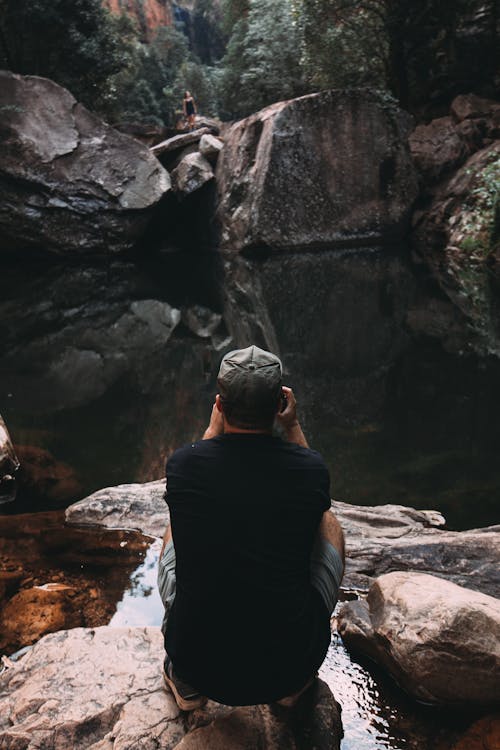 Back view of anonymous male traveler in casual clothes and cap sitting on haunches in gorge and taking photo of distant woman standing on other side of pond
