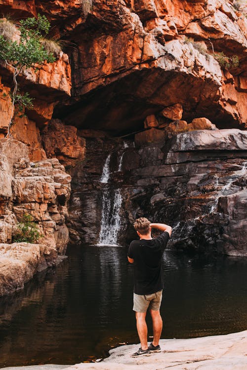 Free Back view of unrecognizable male traveler in casual outfit standing on lake shore and taking photo of picturesque waterfall streaming through rocky cliff in sunlight Stock Photo