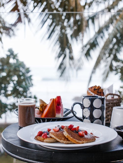 Appetizing pancakes with berries served on table with cold coffee drink and fresh tropical fruits during breakfast on terrace of hotel