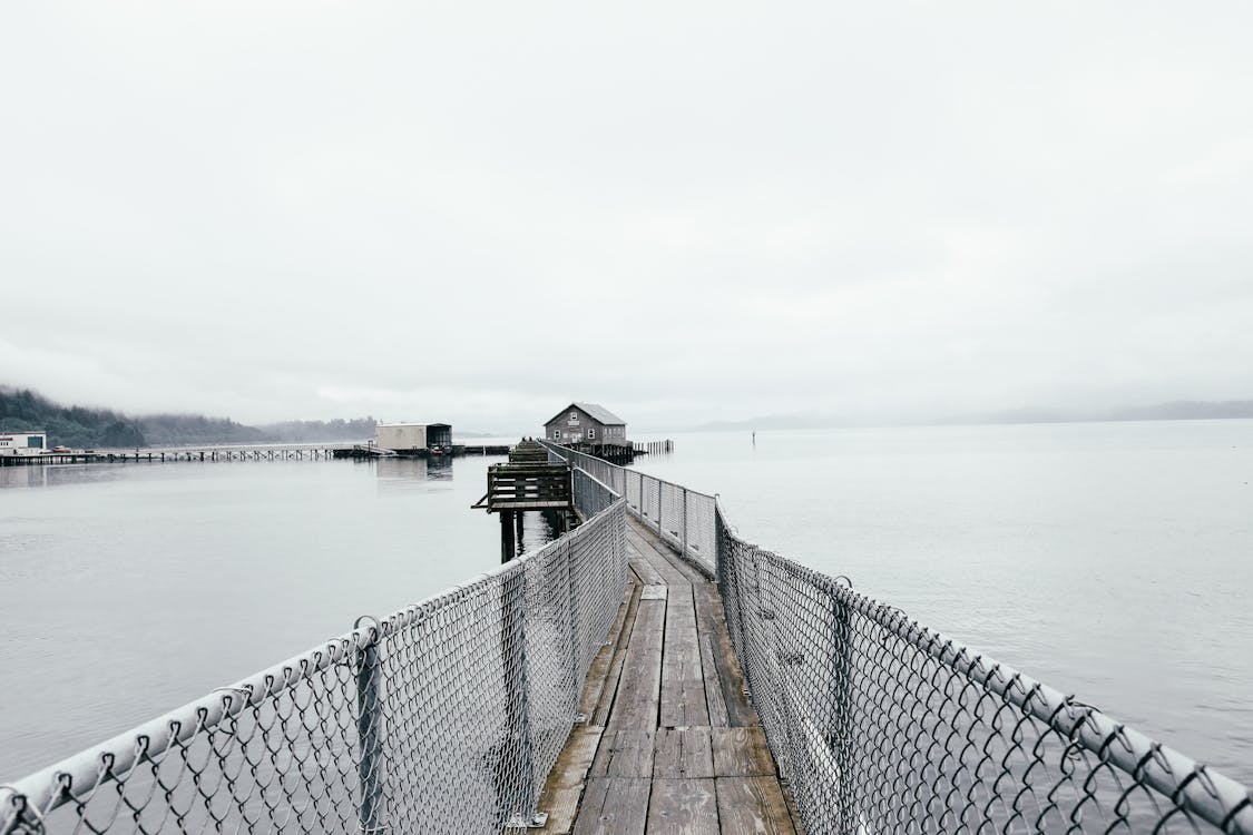 Narrow wooden pier with chain link fence in calm sea near small wooden house against overcast sky in Garibaldi Marina
