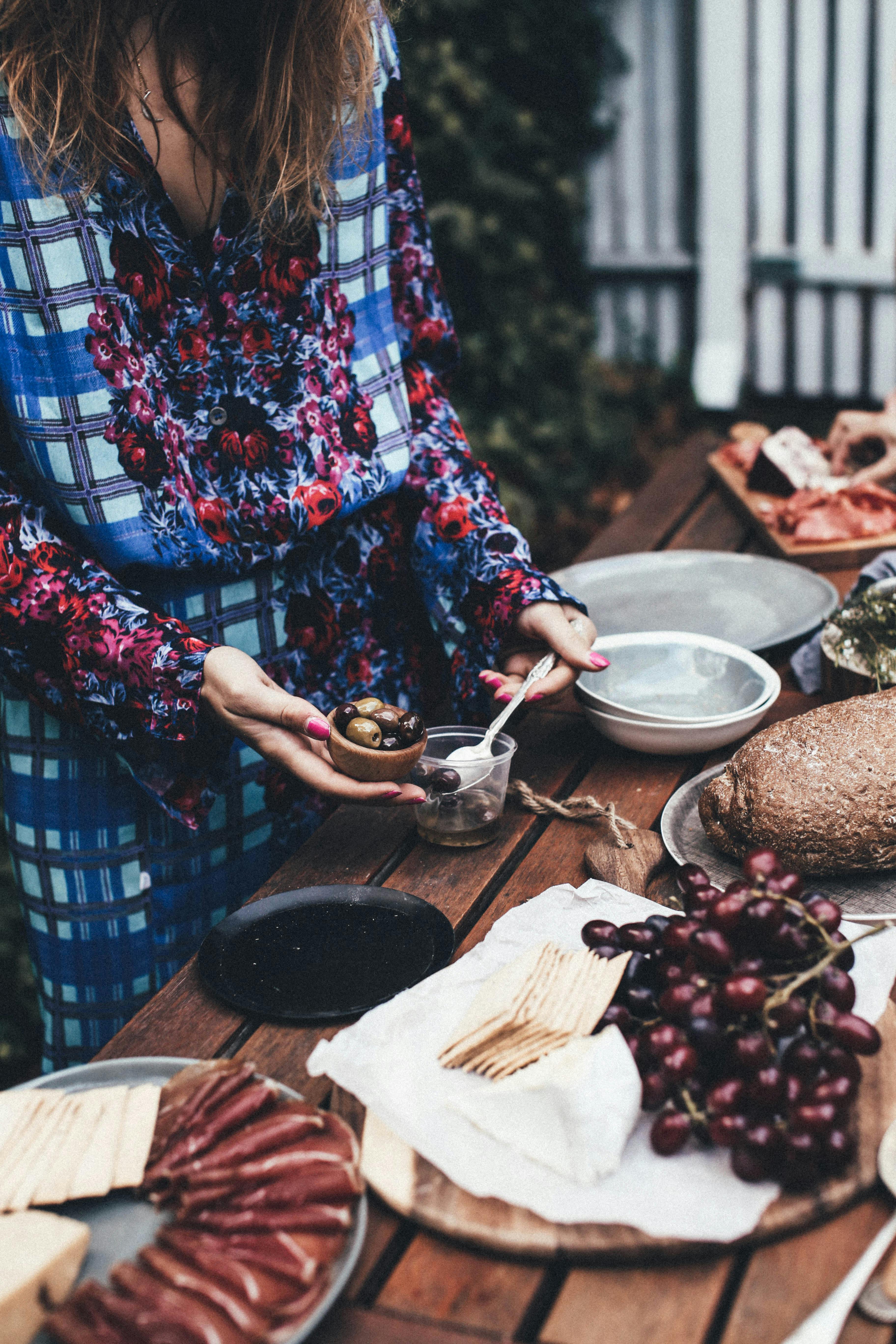 High angle of crop faceless female in stylish dress putting olives into small bowl while serving wooden table with various appetizers during picnic in garden