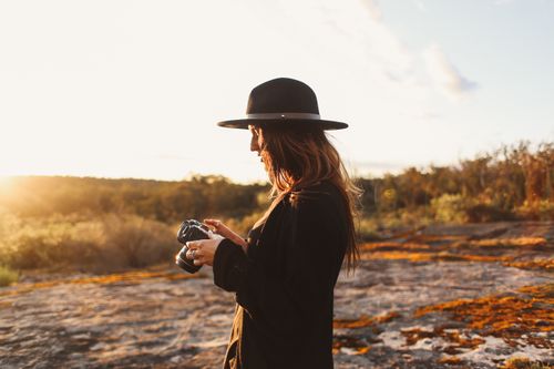 Free Side view of trendy young female traveler with long hair in stylish clothes and hat standing on rocky terrain near dry grassy meadow and setting up photo camera during trip Stock Photo