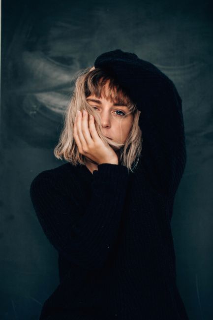 Calm young female model with blond hair in casual clothes touching head and chin and looking at camera against gray wall