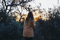 一位长发女游客穿着时尚保暖的衣服，站在郁郁葱葱的绿树旁，在大自然中享受日落时光