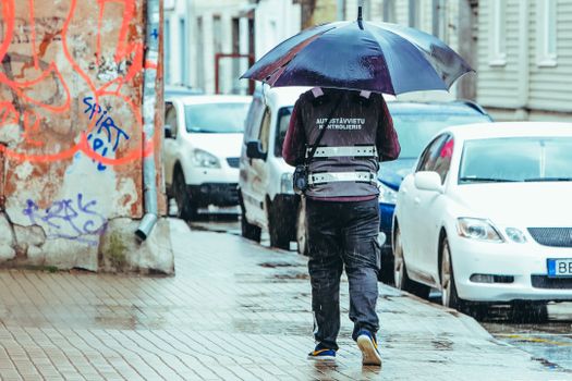 Full body back view of anonymous male scrolling on paved street with umbrella during overcast rainy day