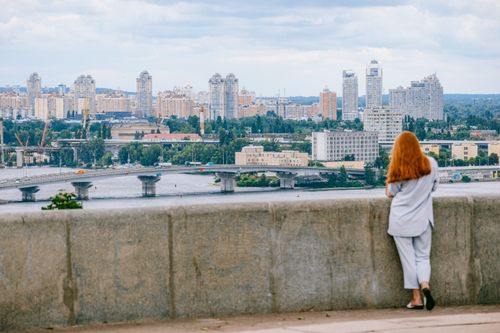 Free A woman with ginger hair overlooks the Kyiv cityscape, featuring high-rise buildings and a river bridge. Stock Photo