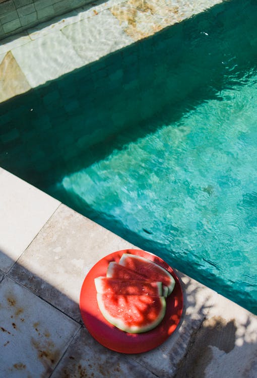 From above of small red slices of watermelon on round plate next to turquoise swimming pool in sunlight