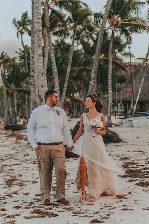 Young content plump man with hand in pocket holding hand of stylish bride with flower bouquet while standing on shore and looking at each other