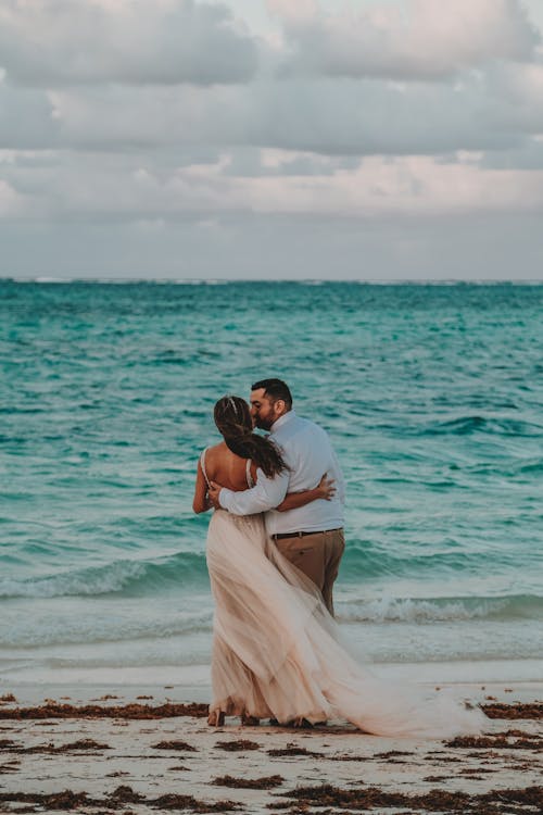 Back view of unrecognizable stylish bride and groom embracing and kissing on sea coast under cloudy sky on wedding day