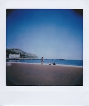 Scenic beach photo featuring tourists, blue sky, and a distant cliff captured in Polaroid style.
