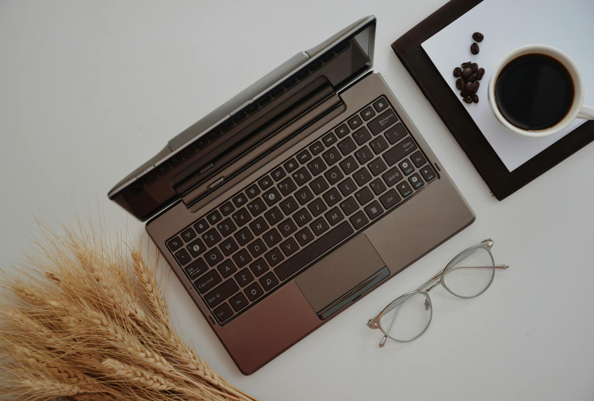 Overhead view of netbook near mug of coffee with golden wheat spikes and eyeglasses on white background
