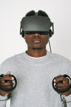 A man wearing VR glasses and gray sweater interacts with virtual reality controllers on a white background.