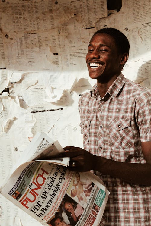 Smiling African American male in checkered shirt with closed newspaper in hand standing near shabby board with papers while looking away