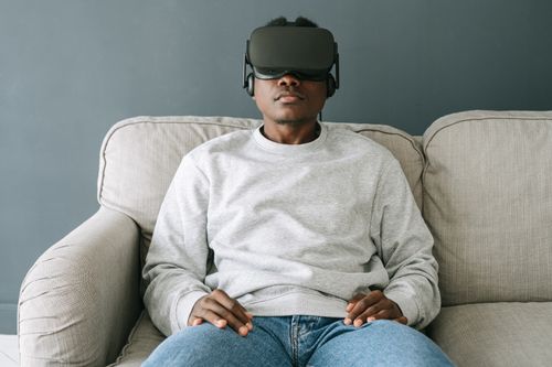 Free Man sitting on a couch wearing VR goggles, exploring virtual reality in a cozy room. Stock Photo