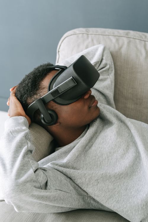 Free A man in casual attire relaxing on a sofa, immersed in virtual reality using a VR headset. Stock Photo