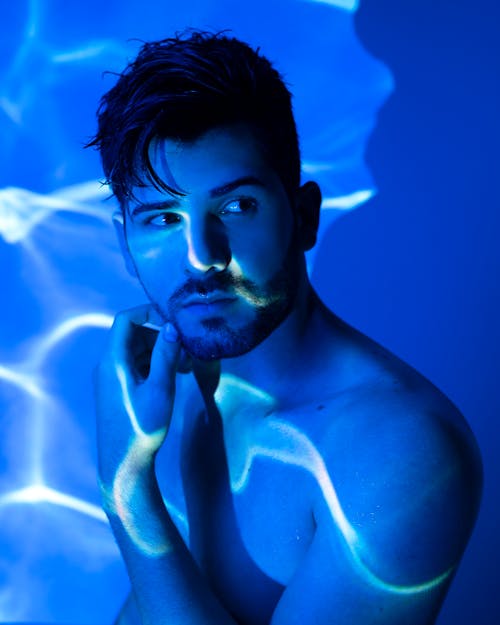 Shirtless male with dark hair standing with hand on chin under glowing neon light and looking away thoughtfully in studio