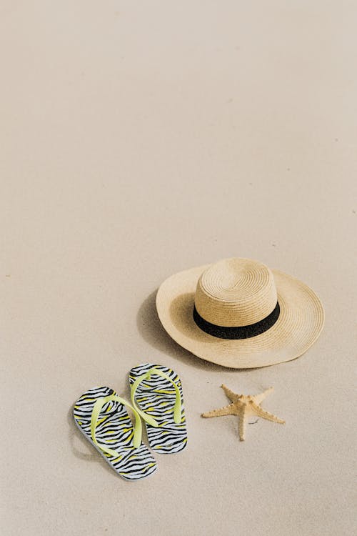 Free Beach Hat and Yellow Strapped Flip Flops Slippers Beside a Starfish on a Sandy Shore Stock Photo