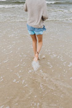 A person enjoys a leisurely walk on the beach in shorts and a sweater, splashing through shallow water.