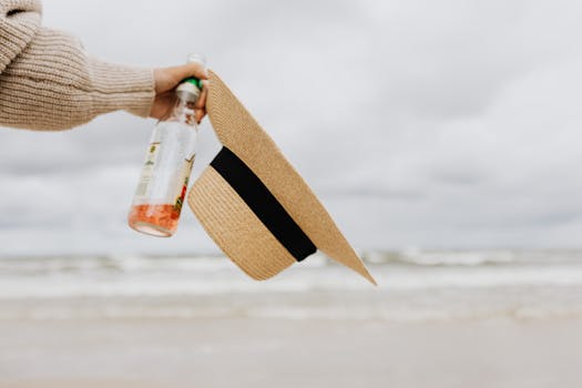 Hand holding a straw hat and beverage on a cloudy beach day. Perfect summer vacation vibe.