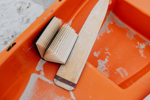 Free An open book and wooden paddle sit inside an orange canoe, captured close-up. Stock Photo