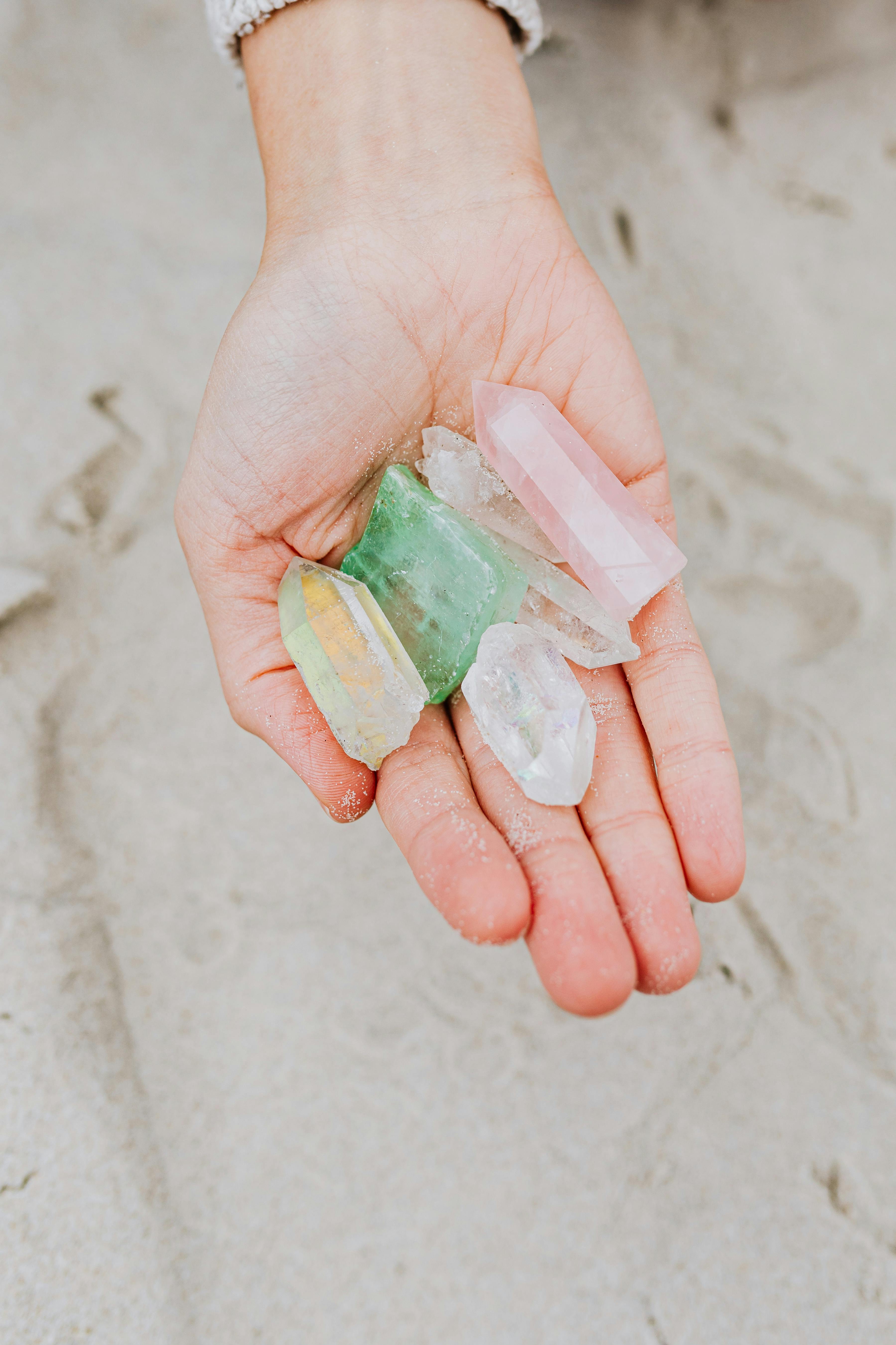 Close-up of a person's hand holding colorful gemstones on a sandy beach shoreline.